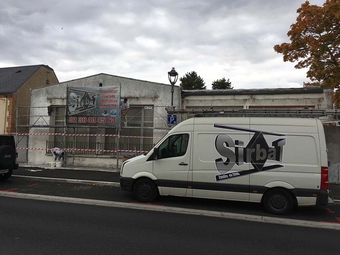 Une camionnette blanche est garée dans une rue de la ville, à côté d'un bâtiment délabré et clôturé, sous un ciel nuageux.