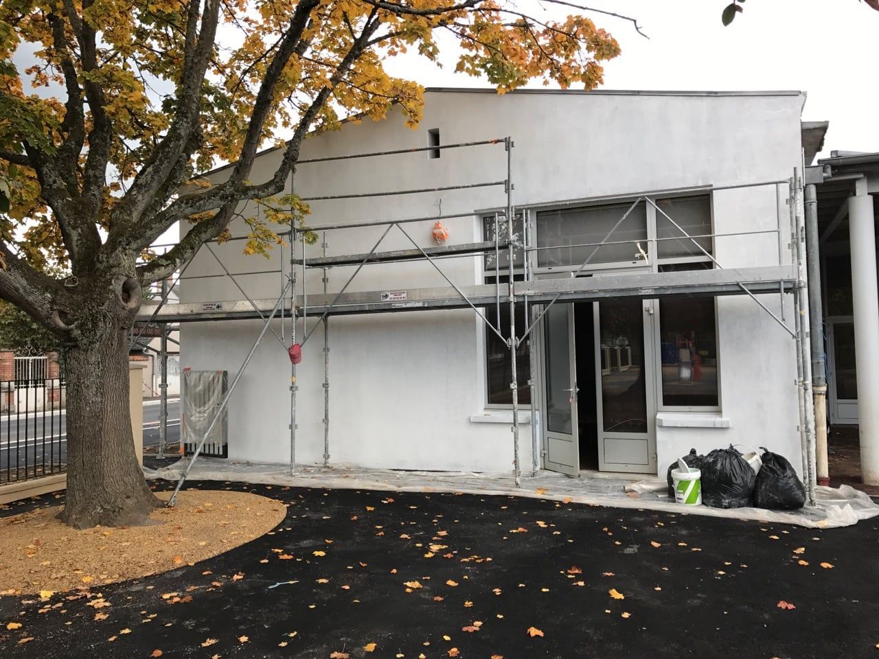 Échafaudages autour d'un bâtiment blanc, avec un arbre devant et des feuilles mortes au sol.