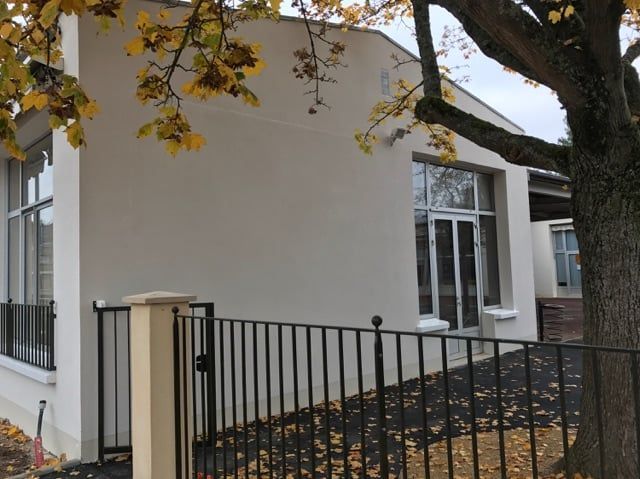 Bâtiment blanc avec de grandes fenêtres et une clôture noire sous les branches d'un arbre d'automne