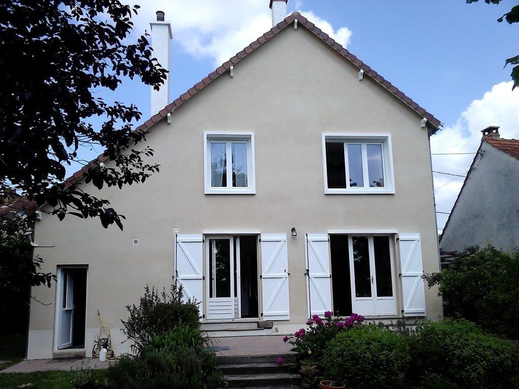 Maison beige à deux étages avec des volets blancs et un petit jardin à l'avant sous un ciel bleu