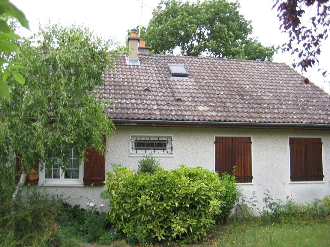 Petite maison blanche au toit de tuiles, aux fenêtres à volets, et des buissons devant, sous les arbres.