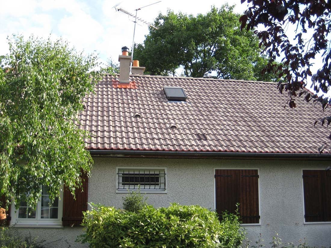 Maison blanche au toit de tuiles brunes, petites fenêtres et arbres devant, sous un ciel nuageux
