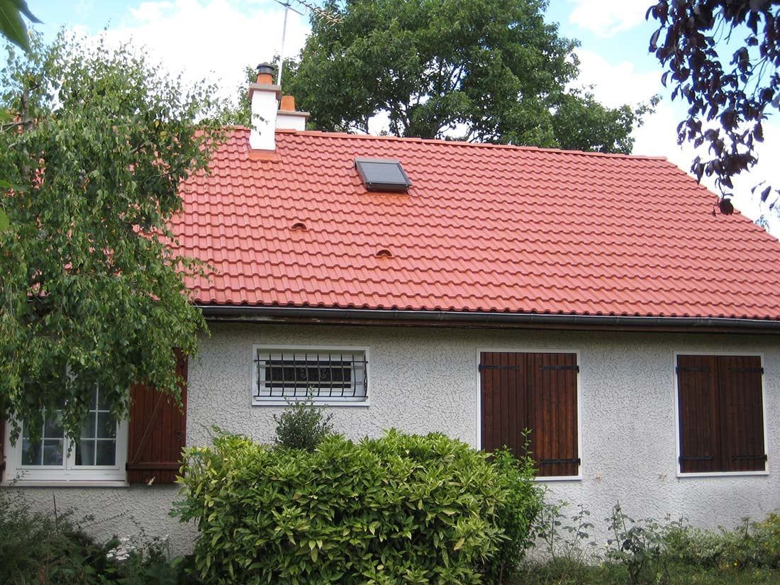 Maison blanche au toit de tuiles rouges, aux volets marrons et aux buissons verts devant, sous les arbres.