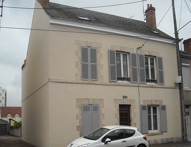 Maison crème de deux étages avec des volets gris et une voiture blanche garée devant, dans une rue résidentielle.