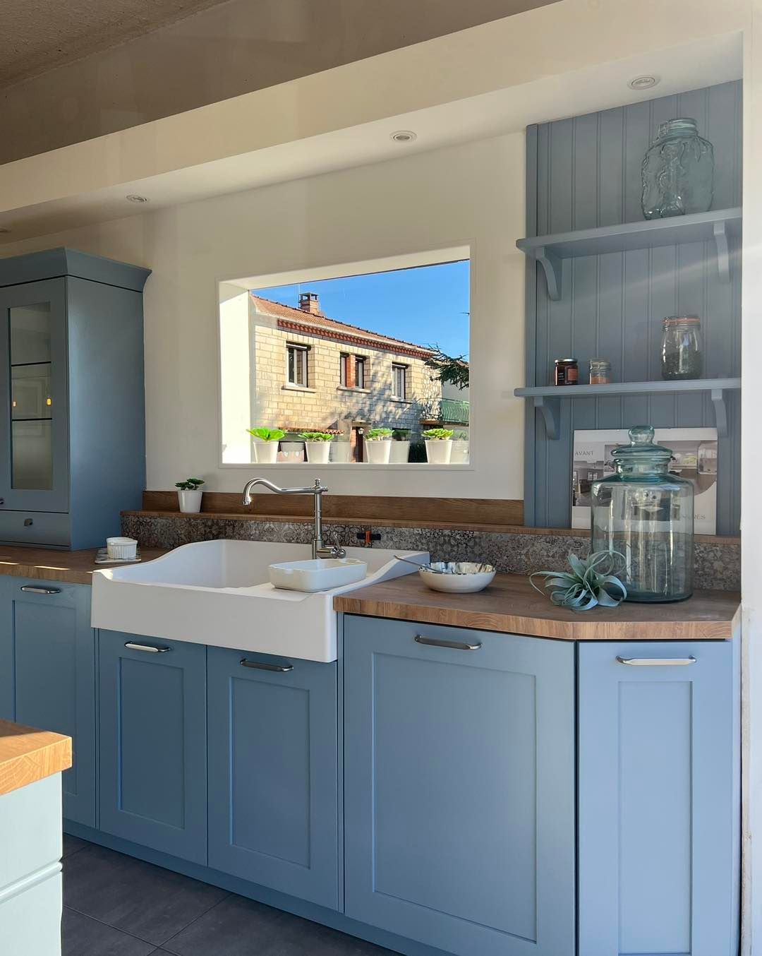 Cuisine style ferme bleue avec évier blanc, comptoir en bois, étagères ouvertes et vue sur des maisons en pierre par la fenêtre