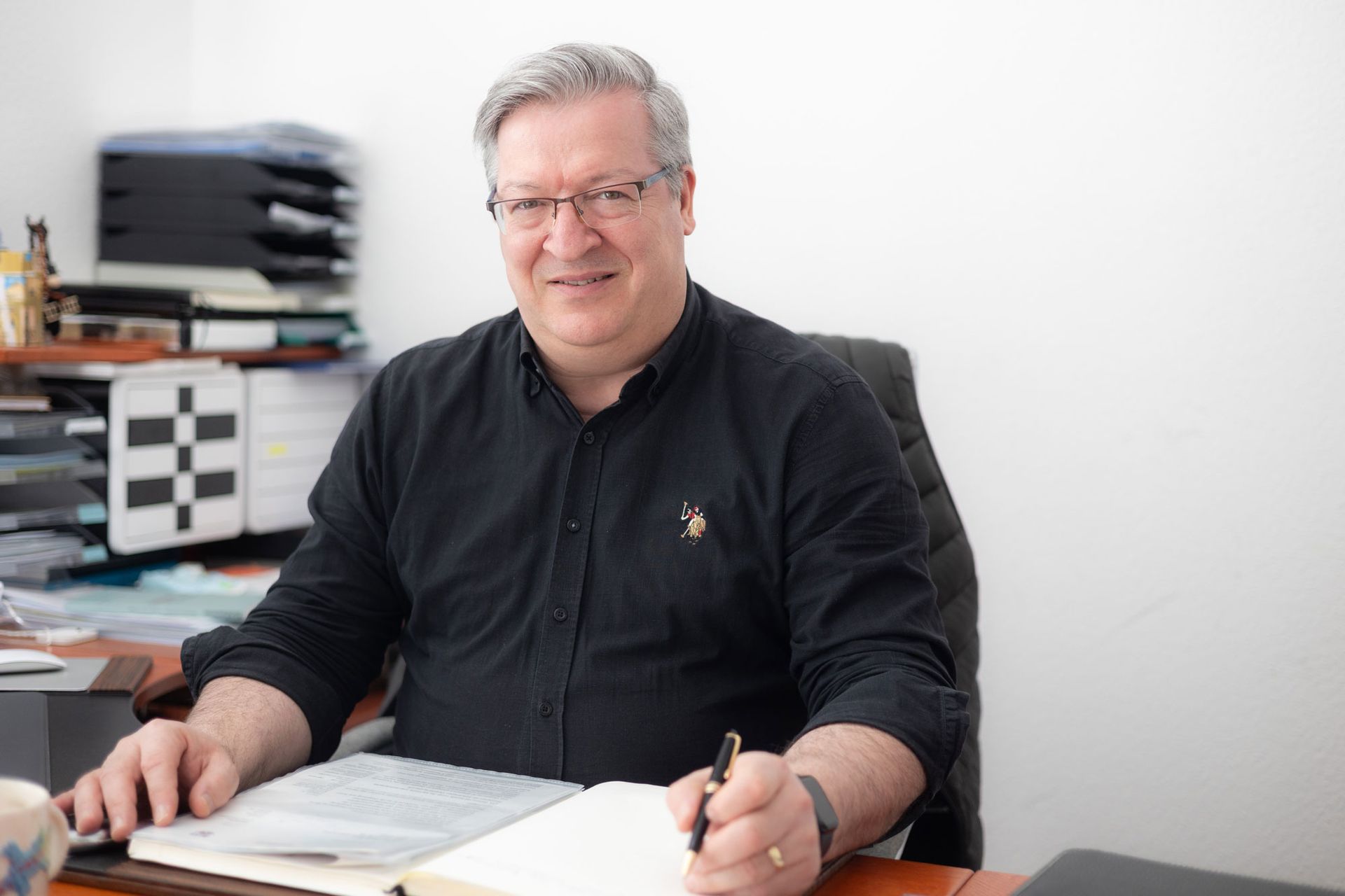 Ein Mann sitzt an einem Schreibtisch mit Papieren und Büromaterialien. Er trägt ein schwarzes Hemd und hält einen Stift in der Hand.