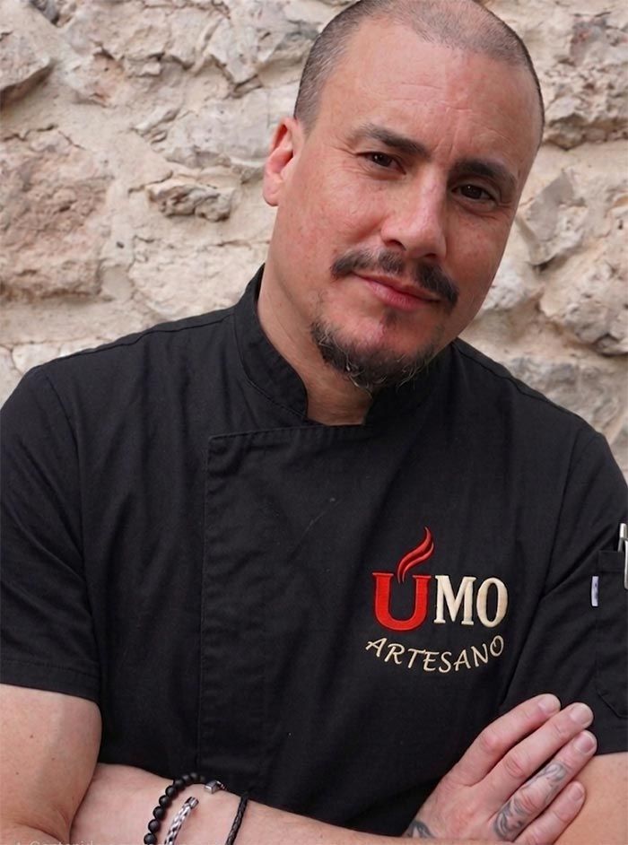 Hombre con los brazos cruzados, vestido con una chaqueta de chef negra y un logotipo rojo, de pie frente a un muro de piedra.