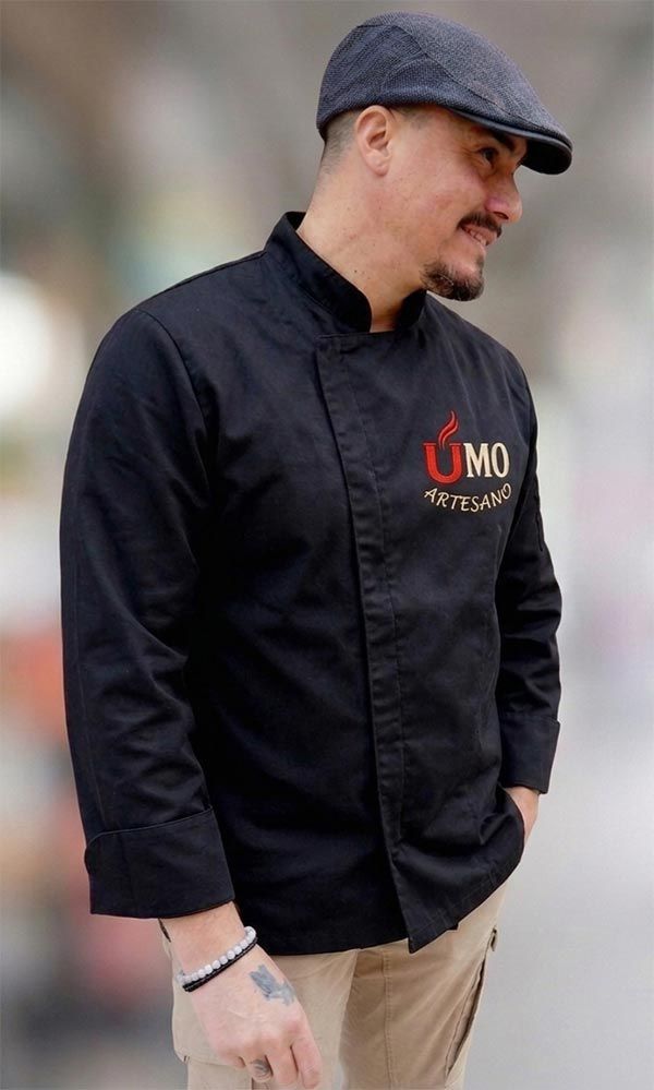 Chef con chaqueta negra y gorra plana, de perfil, con el logotipo “UMO” en el pecho.