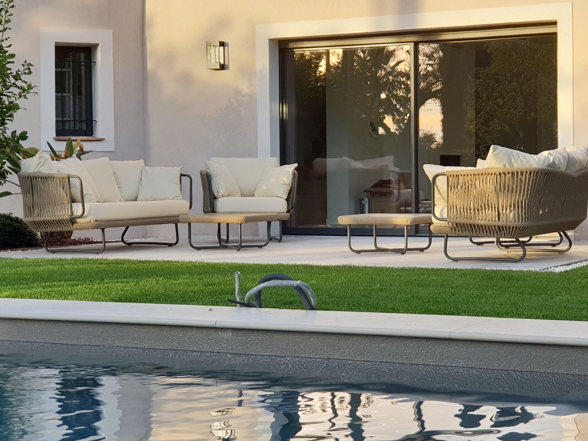 Ensemble de mobilier de jardin près d'une piscine. Canapés beiges à côtés tressés, tables basses et portes coulissantes en verre.