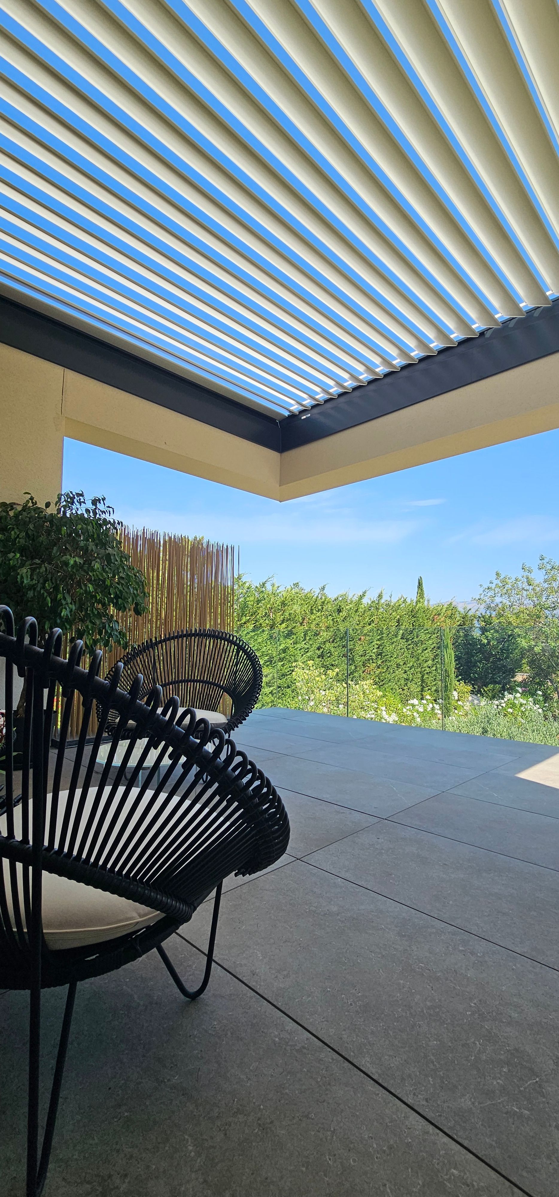 Terrasse extérieure avec chaises en osier noir sous une pergola à claire-voie, ciel bleu visible.