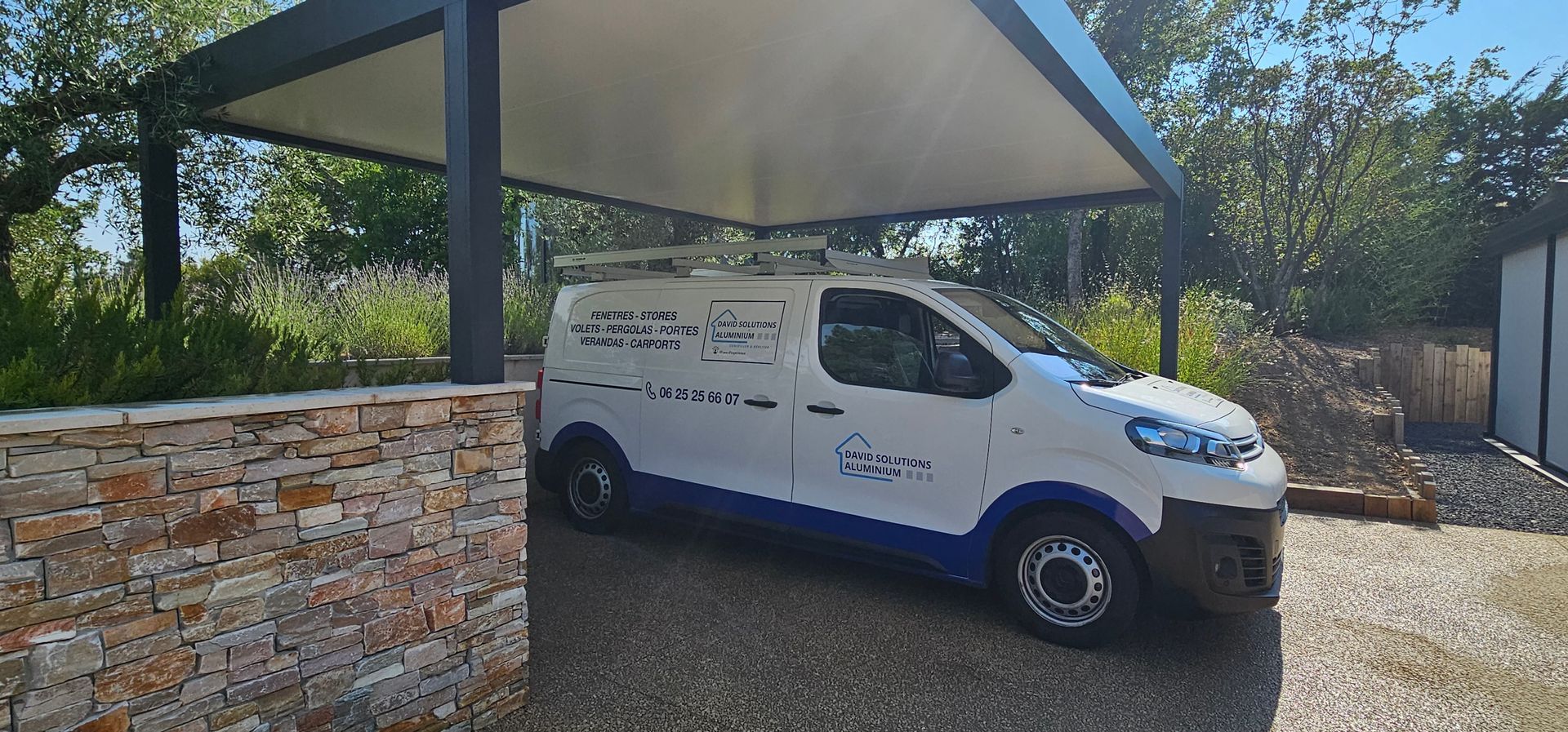 Une camionnette de professionnels est garée sous un carport, avec un muret en pierre au premier plan.