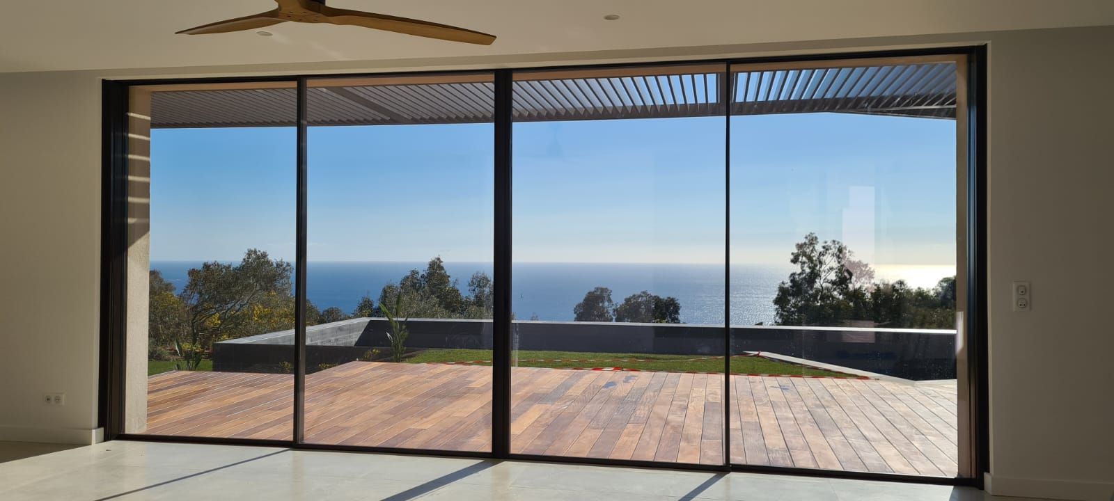 Grandes portes coulissantes en verre offrant une vue sur l'océan, les arbres et le ciel. Terrasse en bois.