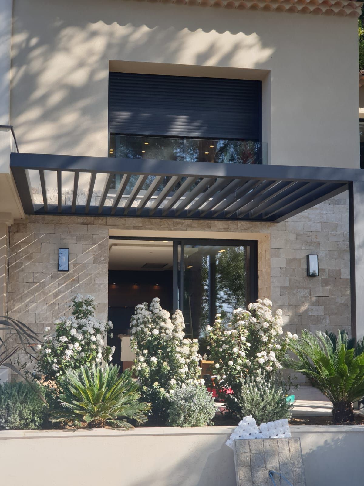 Maison avec pergola, jardin et volets fermés. Murs beiges, pergola grise.