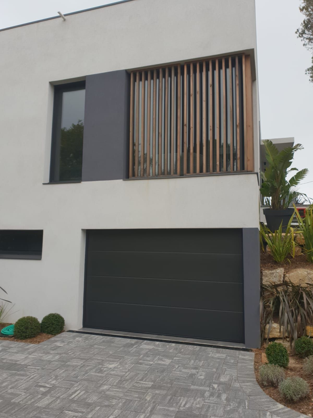 Maison blanche avec porte de garage grise, fenêtre et détails en lattes de bois. Allée pavée et aménagement paysager.