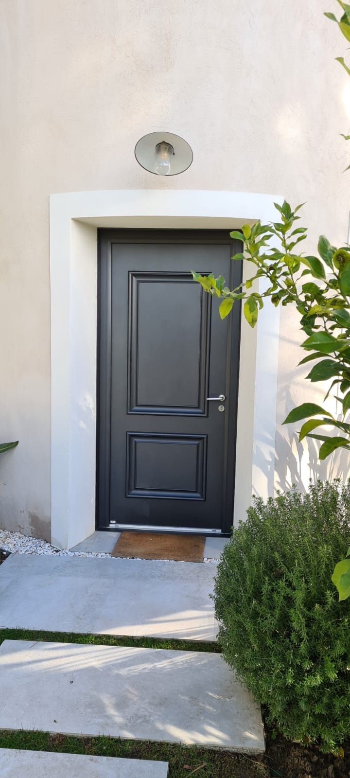 Une porte d'entrée noire avec des encadrements blancs, un petit tapis et un buisson.