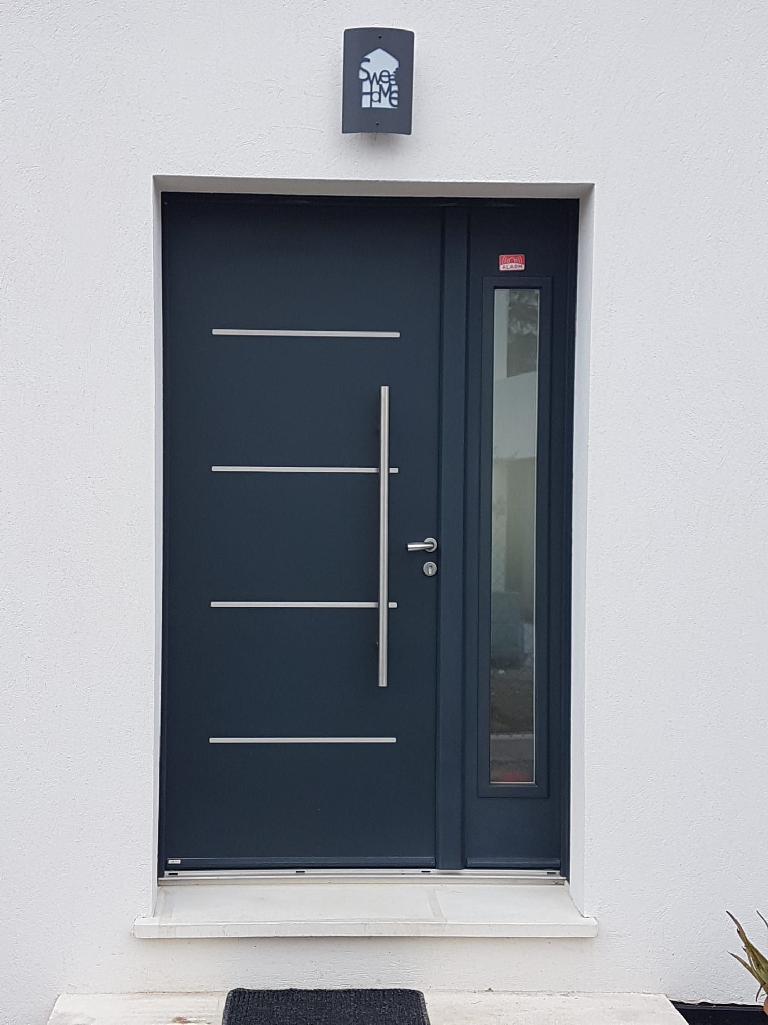 Porte d'entrée gris foncé avec poignée verticale, panneau de verre et trois lignes horizontales argentées.