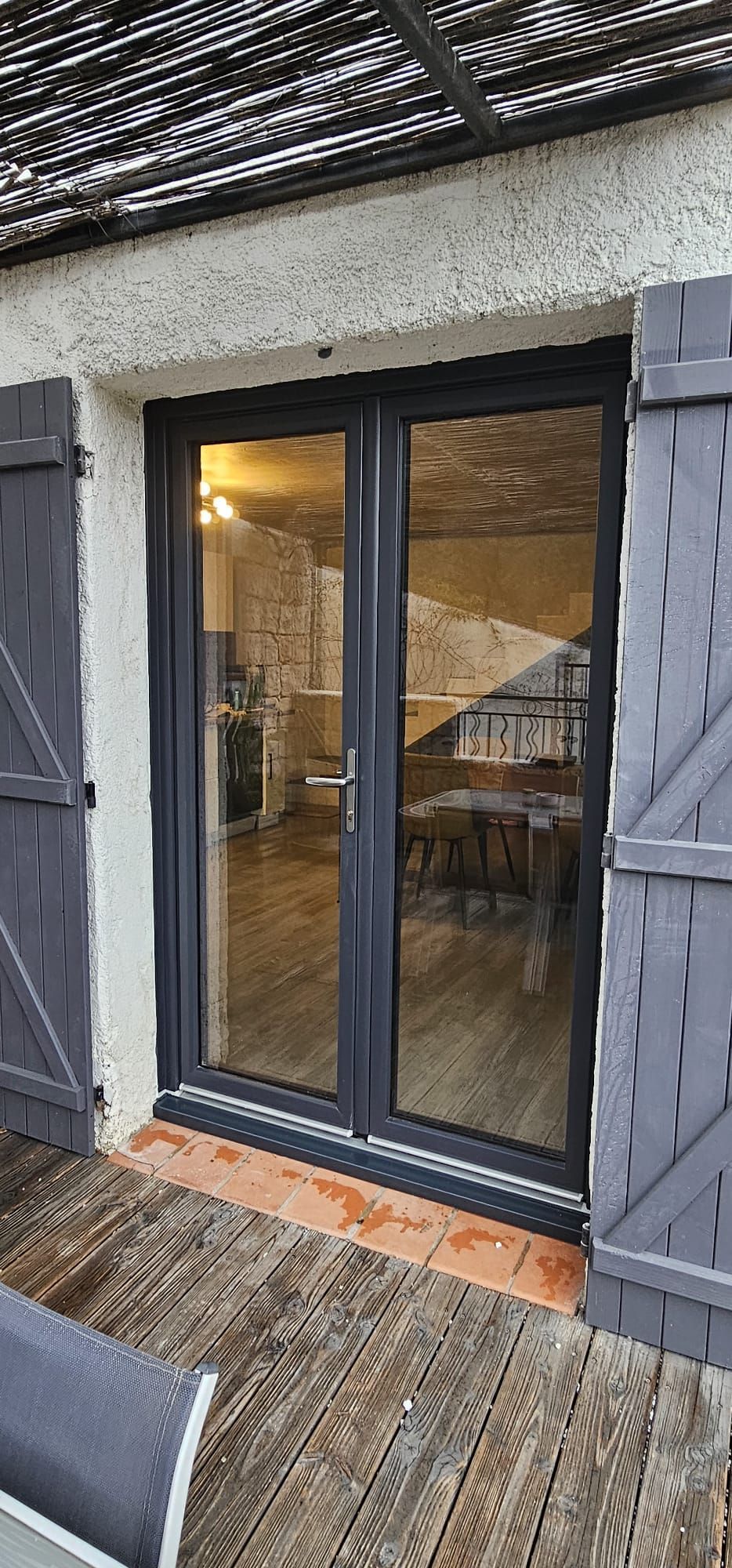 Une porte vitrée à cadre sombre en PVC. Terrasse en bois et façade en pierre.