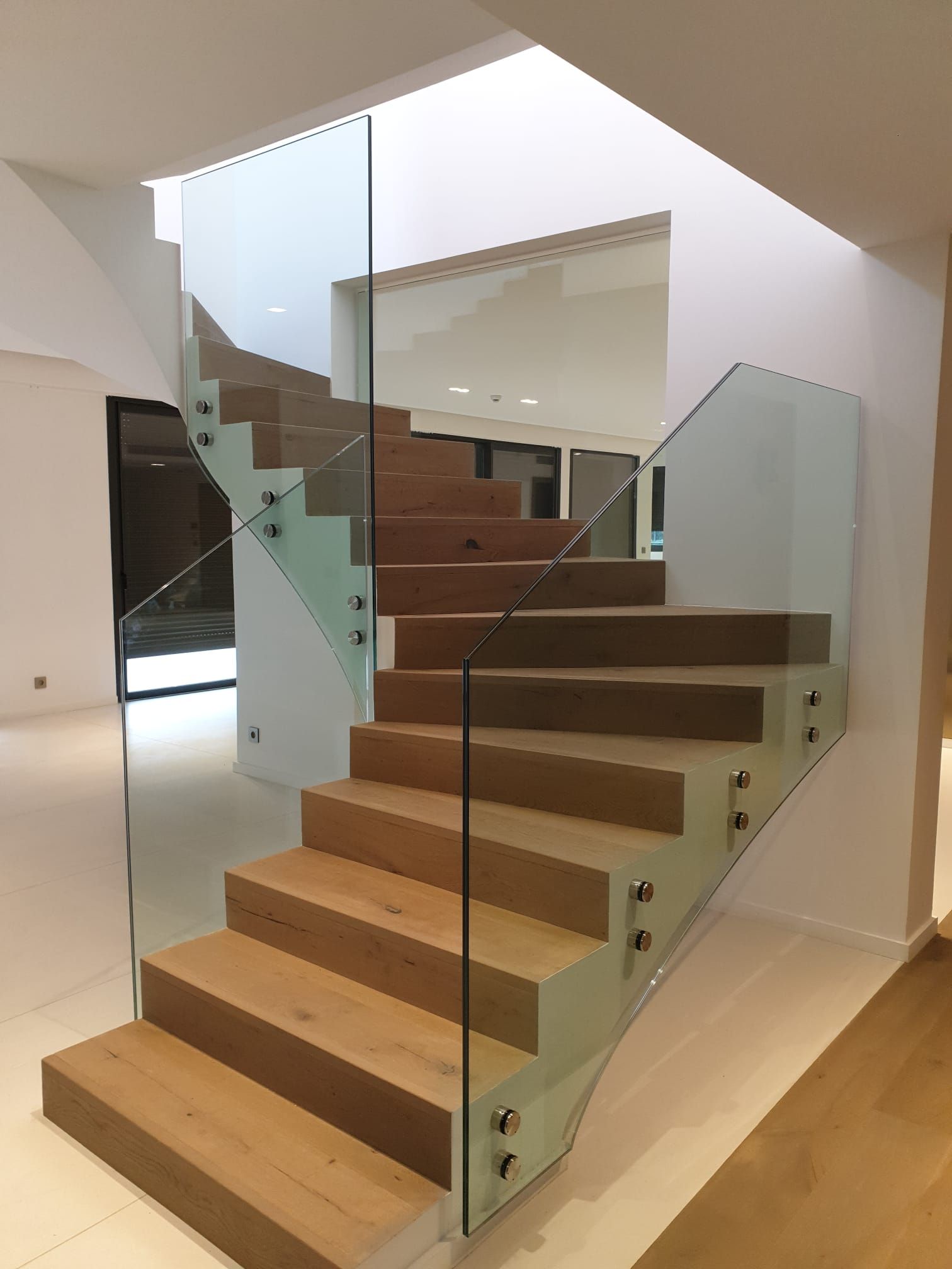 Escalier en bois avec rampe en verre. Murs et sol blancs.