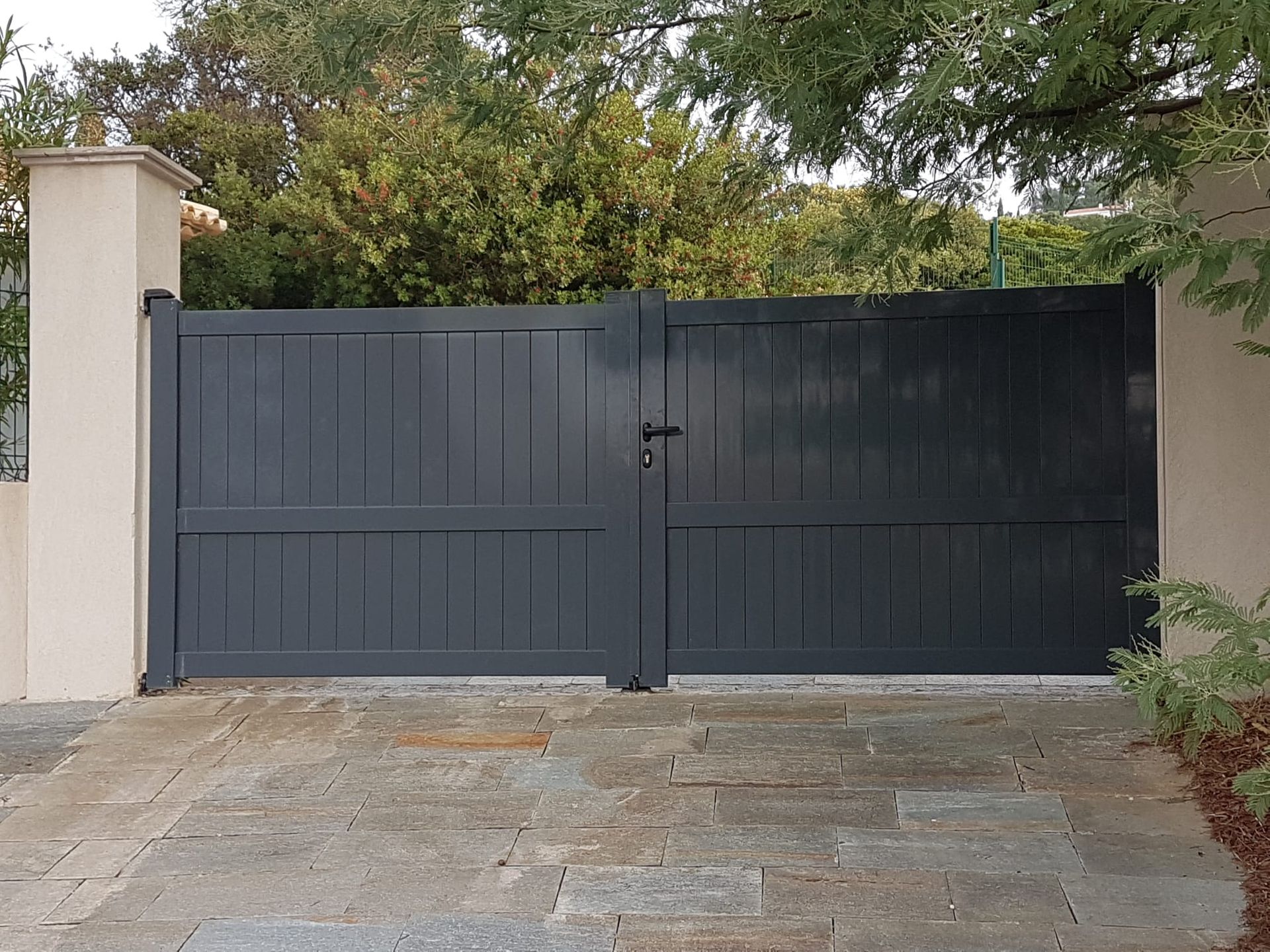 Portail double gris, à planches verticales, centré sur une allée en pierre, flanqué de piliers et de verdure.