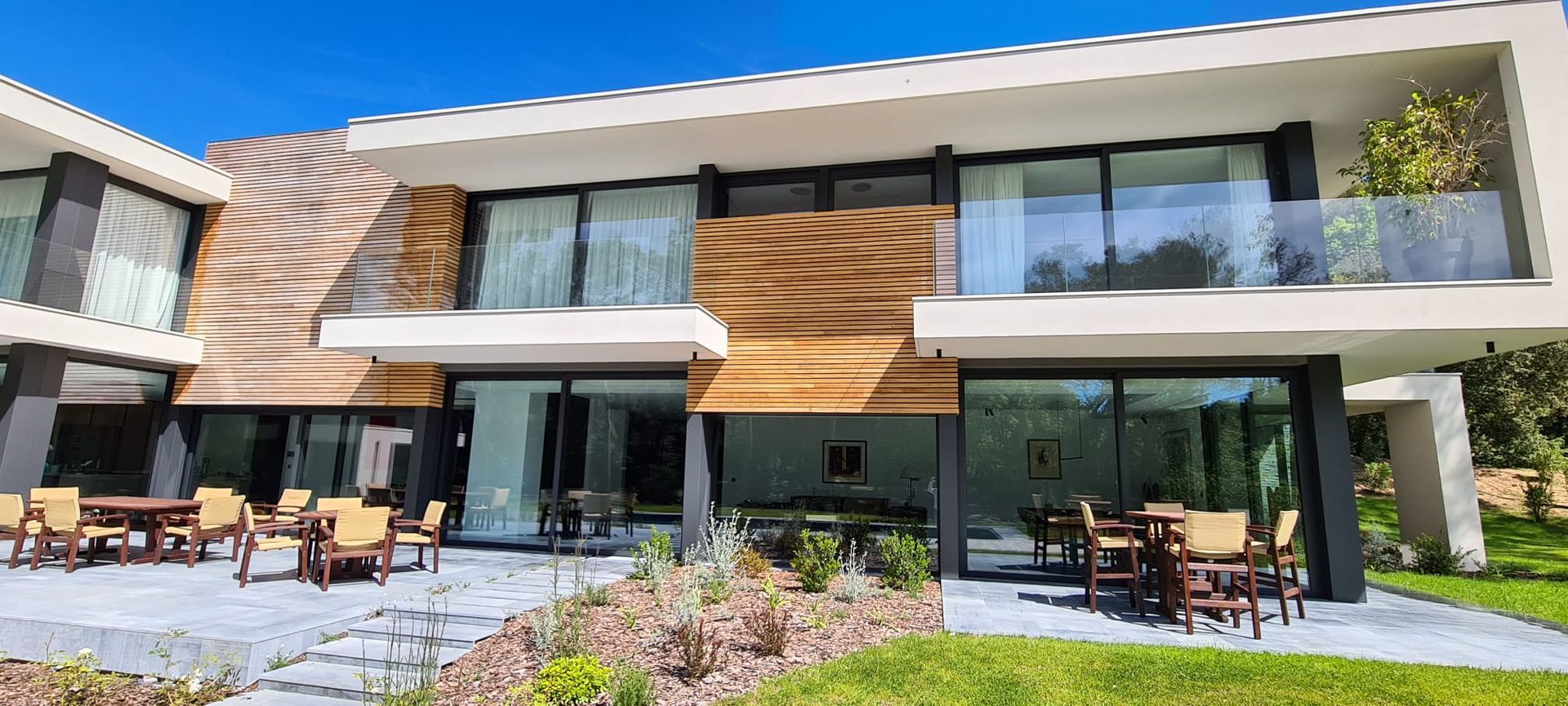 Bâtiment de deux étages avec de grandes fenêtres et des éléments en bois, coin salon extérieur sur une terrasse pavée.