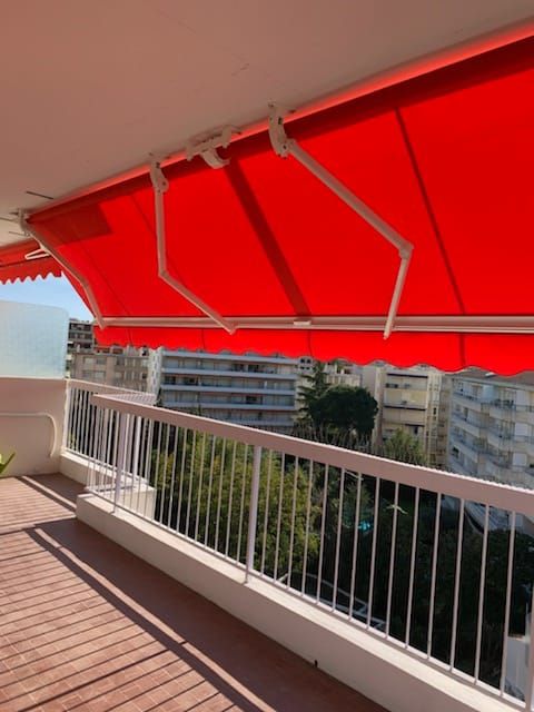 Un store rouge est déployé sur un balcon donnant sur des bâtiments et des arbres.