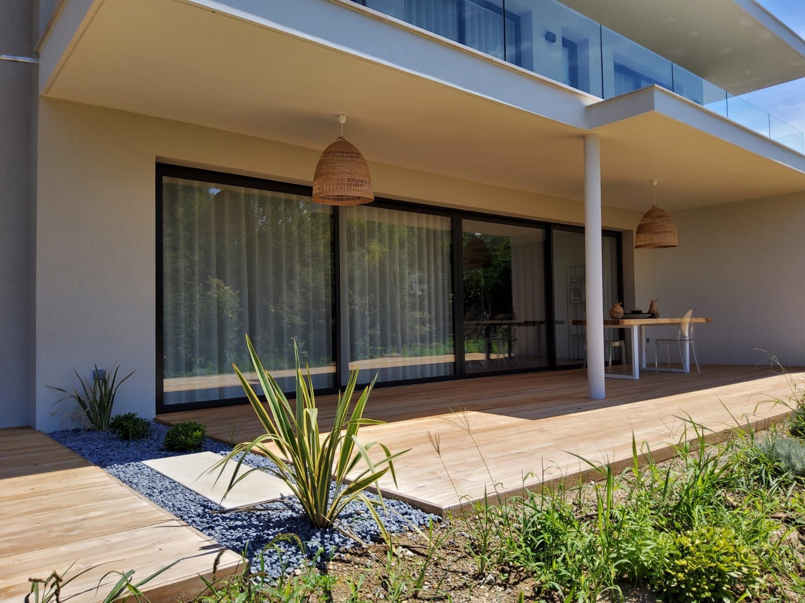 Extérieur d'une maison avec une terrasse en bois, de grandes fenêtres et des lampes suspendues en osier.