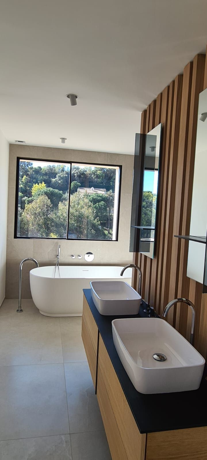 Salle de bains avec baignoire, deux lavabos et une fenêtre donnant sur les arbres.