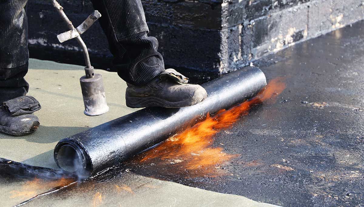 Ein Arbeiter verlegt mit einem Schweißbrenner eine schwarze Dachmembran auf einem Flachdach; orangefarbene Flammen sind sichtbar.