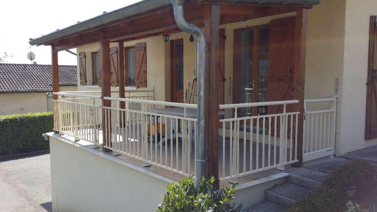 Maison beige avec porche, balustrade blanche, volets en bois et haie verte.