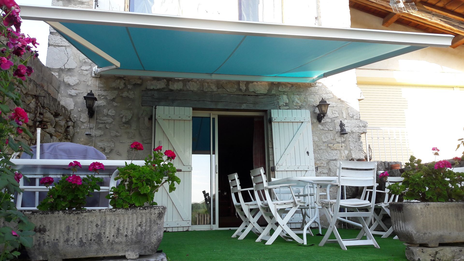 Un auvent bleu ombrage une terrasse meublée de meubles blancs, devant un bâtiment en pierre aux portes ouvertes.