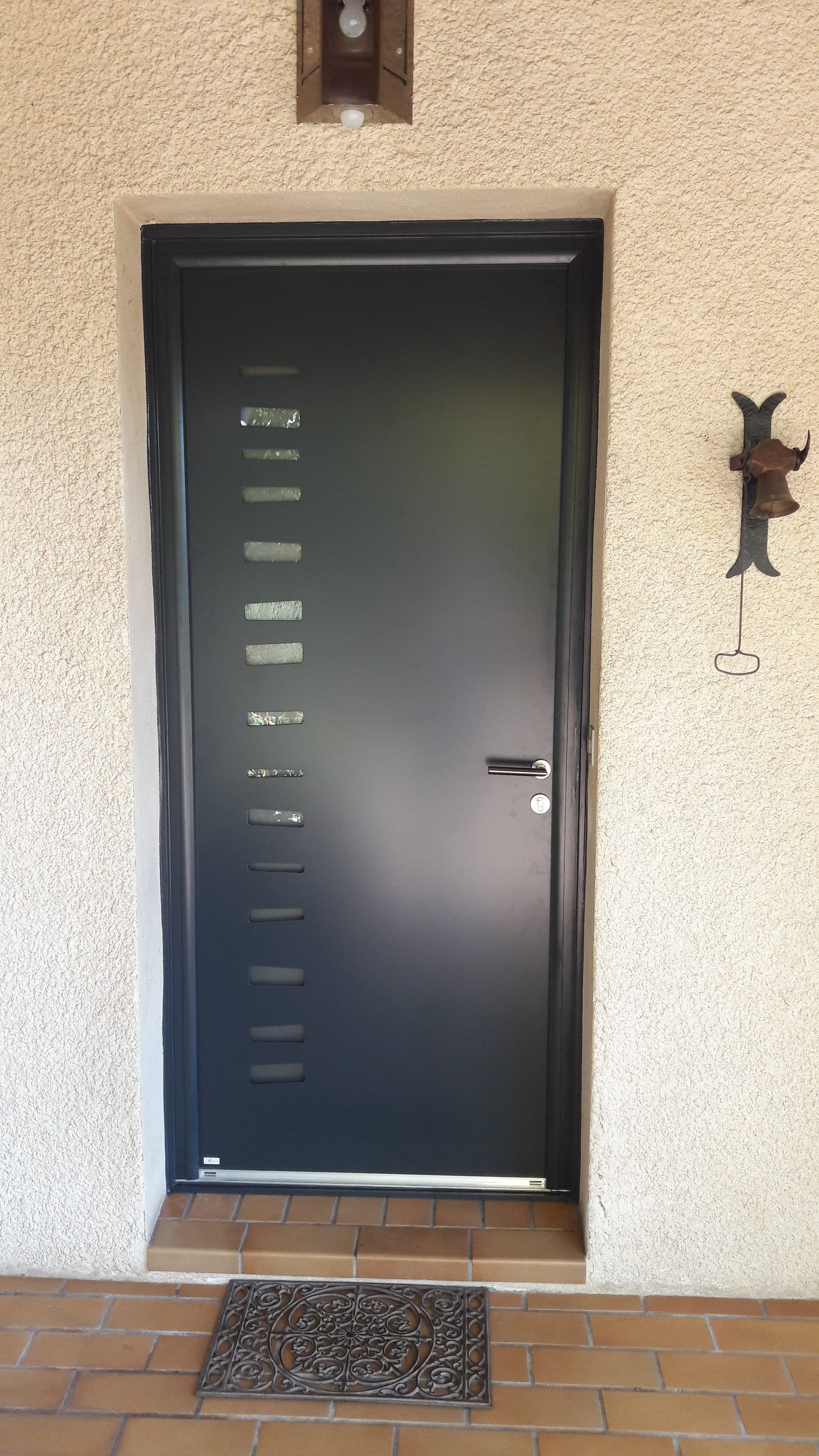 Porte gris foncé avec vitres rectangulaires, paillasson, sonnette sur un mur texturé.