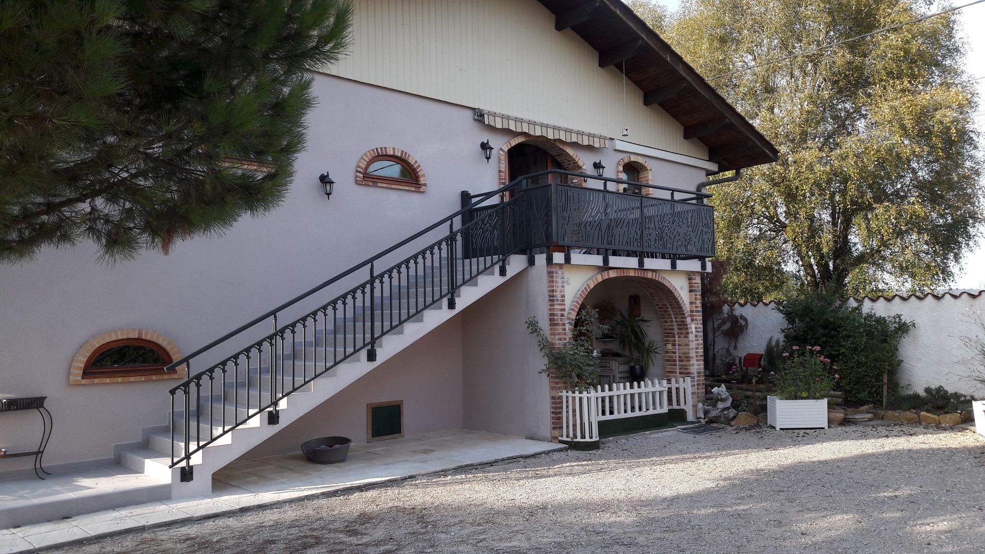 Extérieur d'un bâtiment clair avec un escalier menant à un balcon. Rambardes noires et détails arqués.