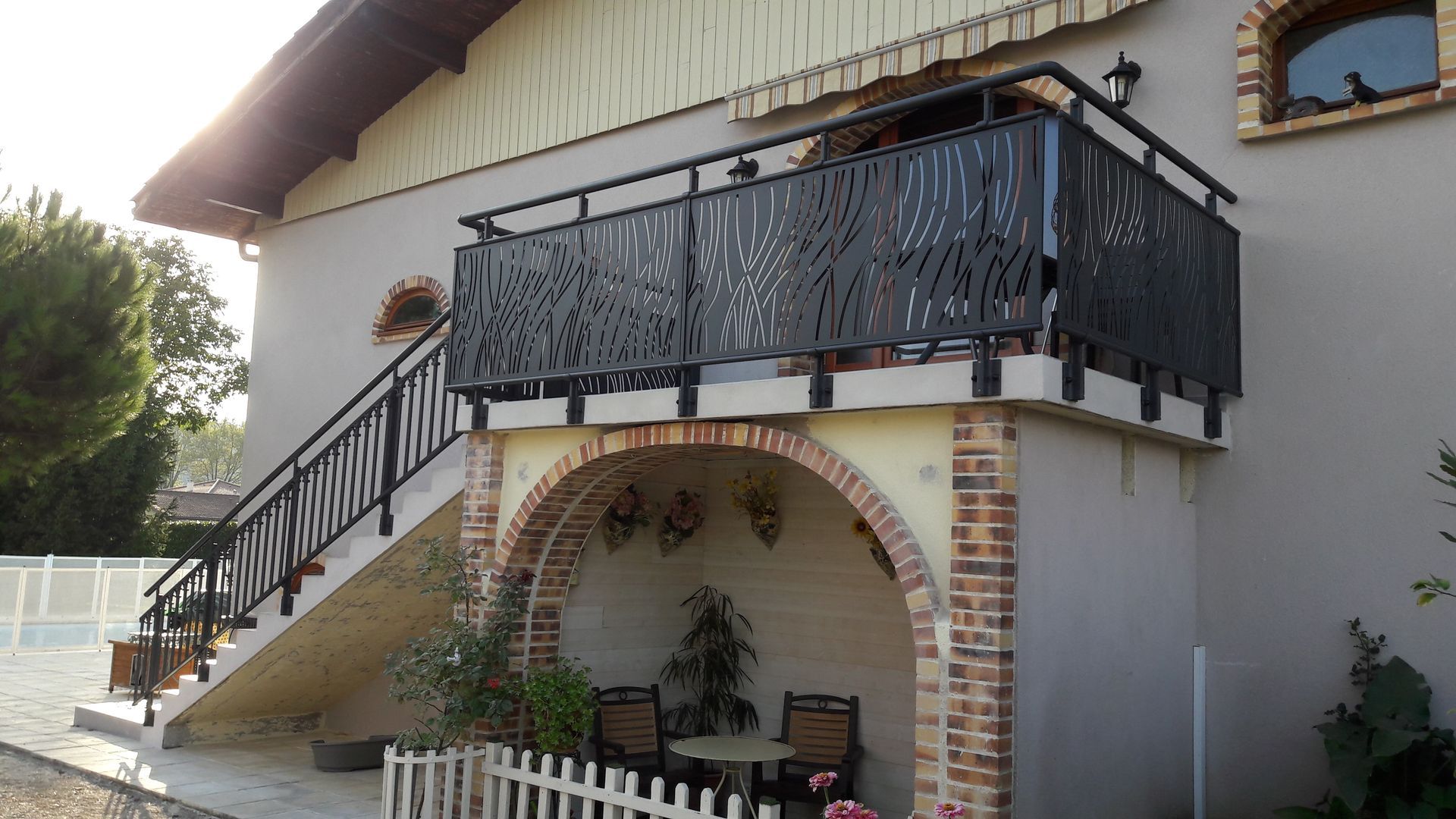 Maison à deux étages avec balcon et escalier en métal noir, arche en briques et petit coin salon.