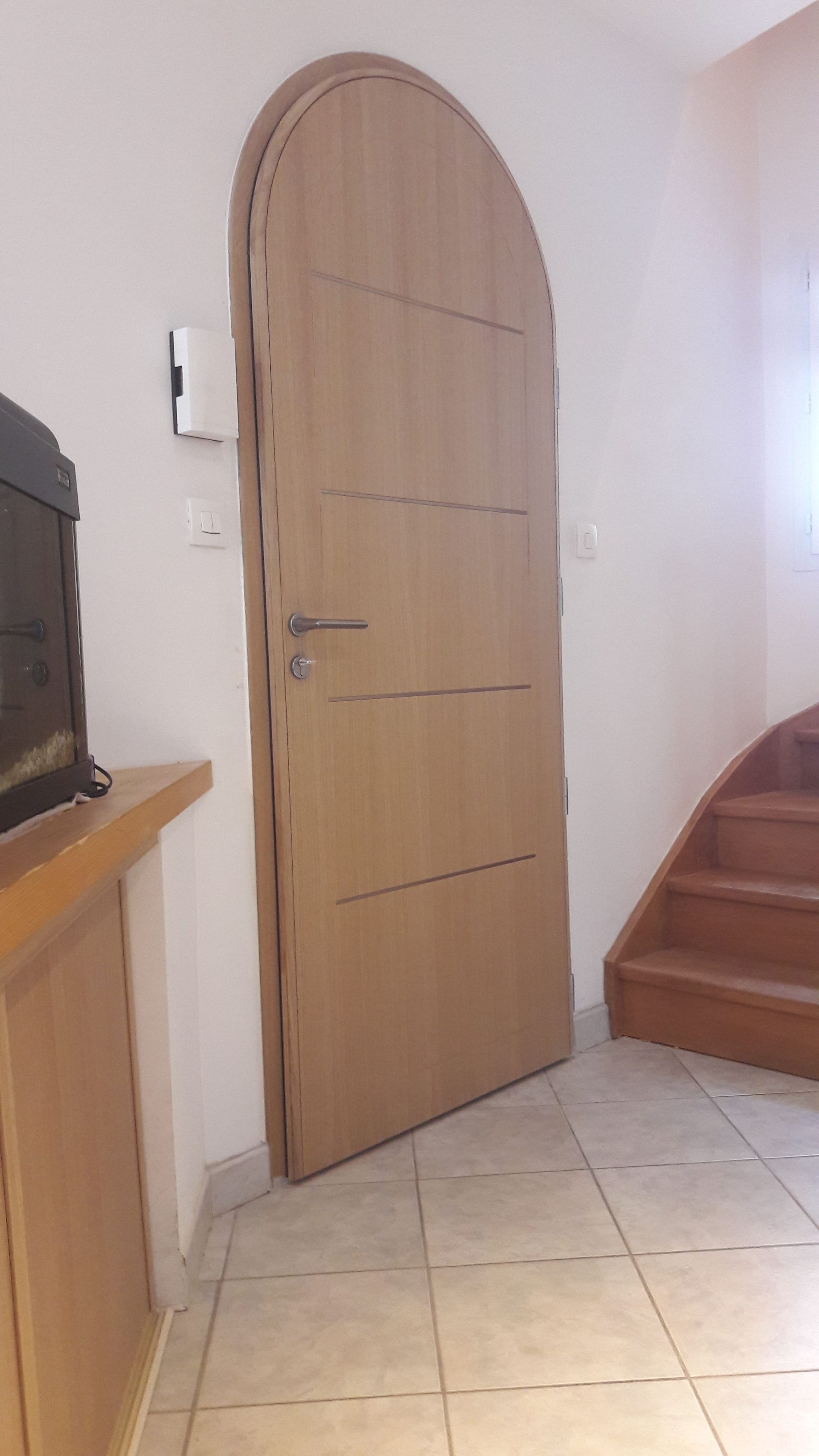 Porte cintrée en bois dans un couloir, à côté d'un escalier et d'une console sur laquelle se trouve un objet sombre.