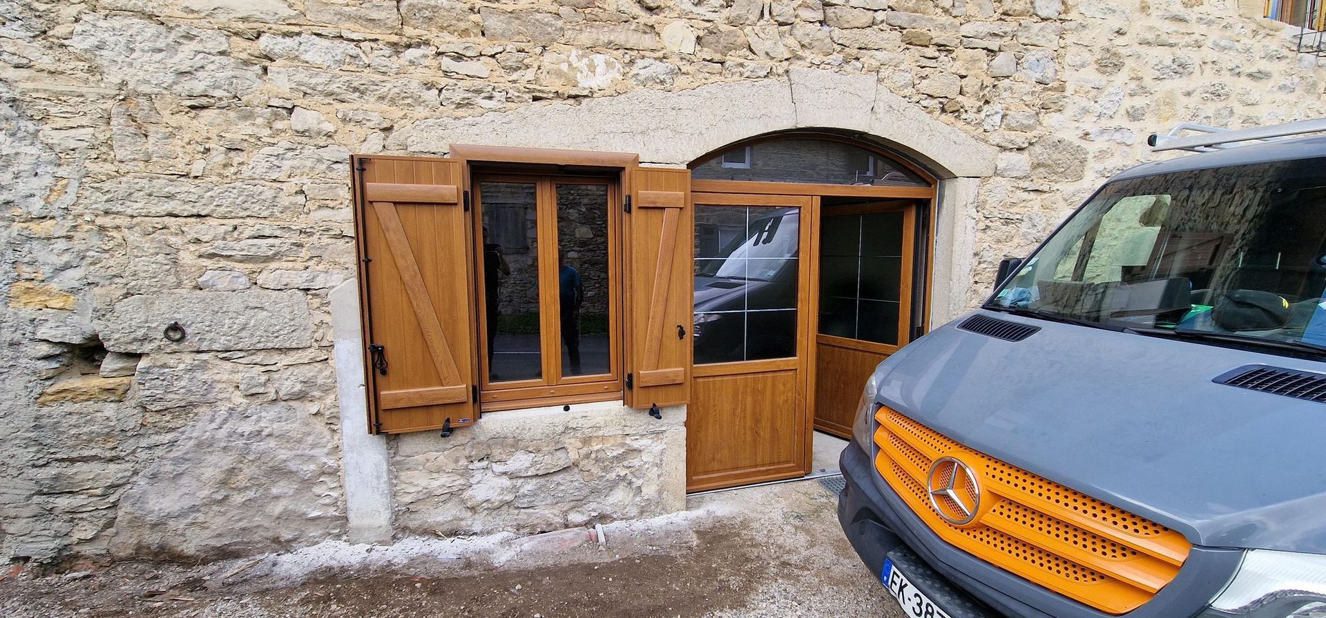 Un bâtiment en pierre avec une porte et des volets en bois, une camionnette grise garée à proximité.