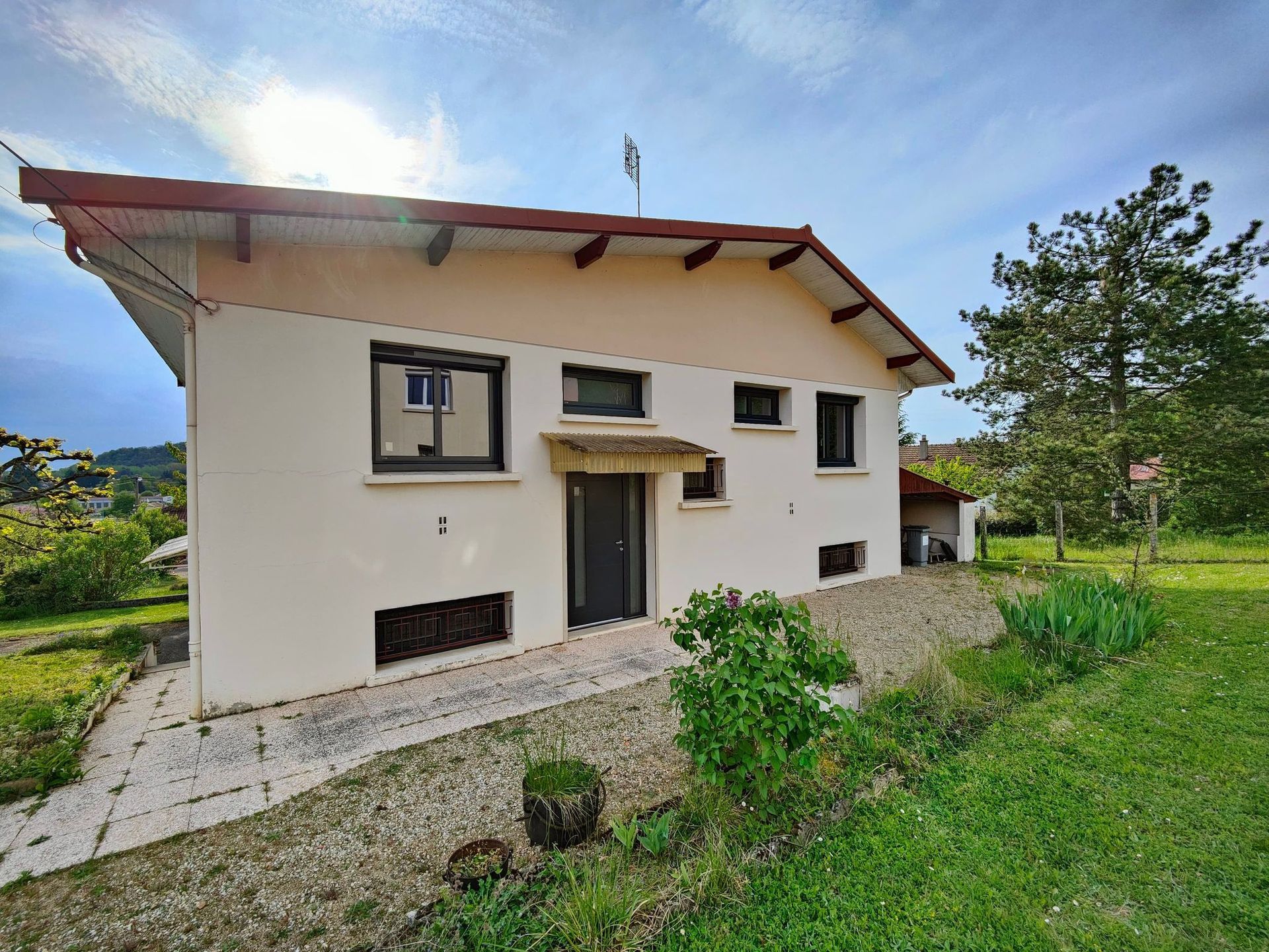 Maison beige au toit rouge, portes et fenêtres noires, dans un cadre verdoyant sous un ciel bleu.