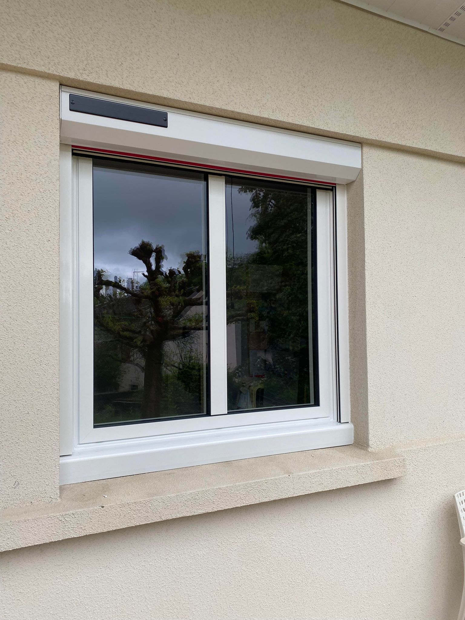 Fenêtre blanche avec volet roulant blanc solaire sur un mur en stuc ; reflets d'arbres dans la vitre.