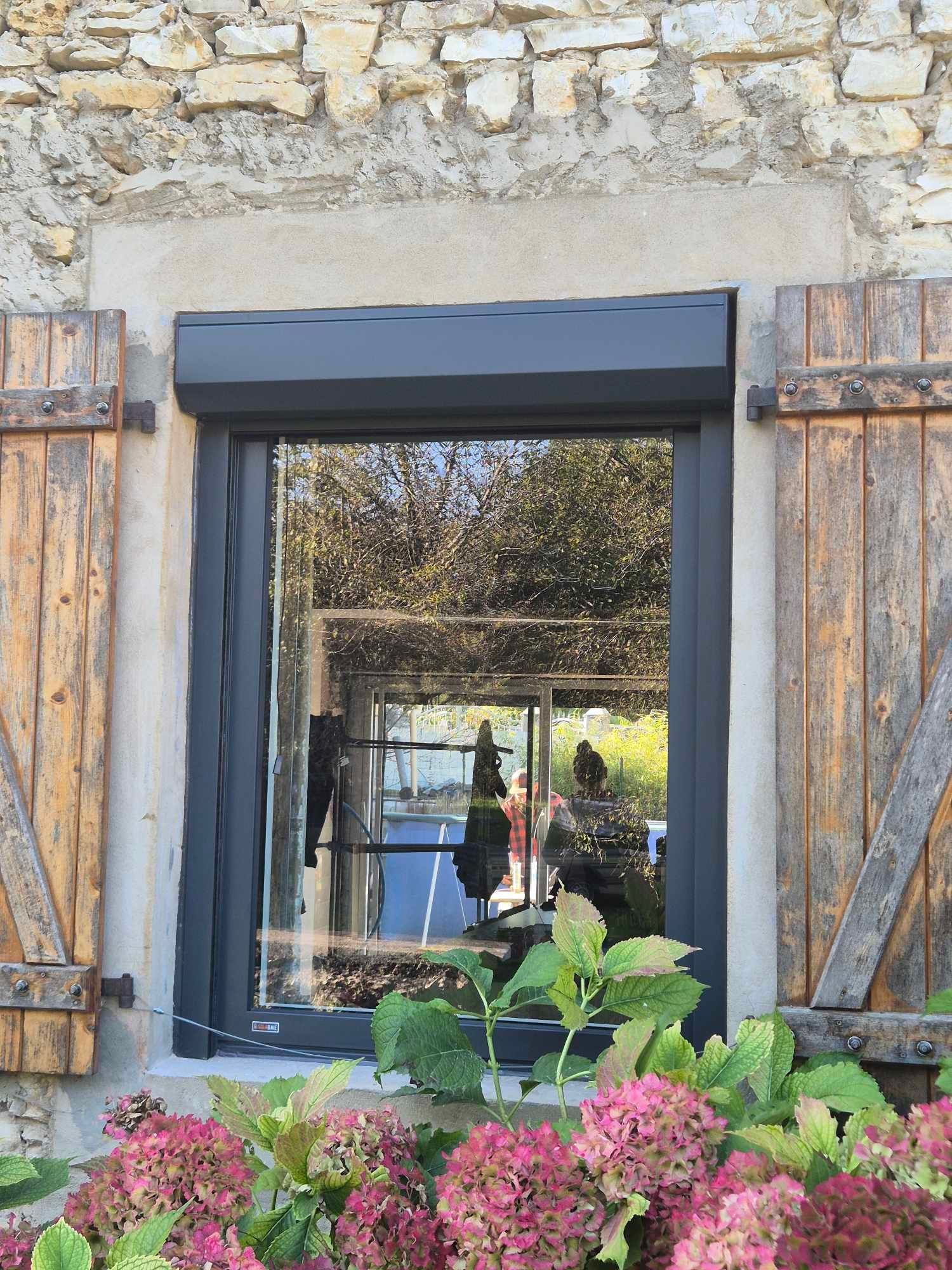 Fenêtre avec volet roulant foncé, volets en bois et fleurs roses devant.