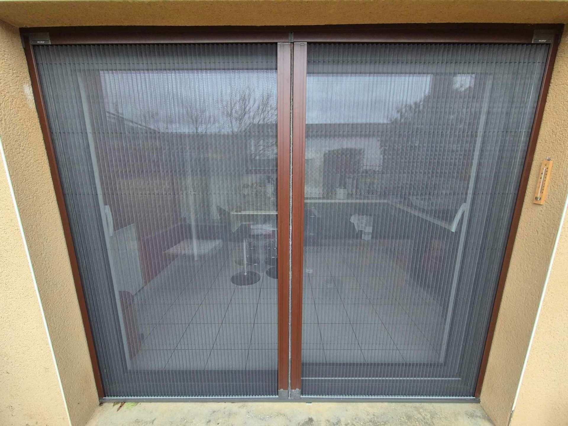 Porte moustiquaire marron, deux panneaux, fermée, masquant la vue sur l'espace extérieur meublé.