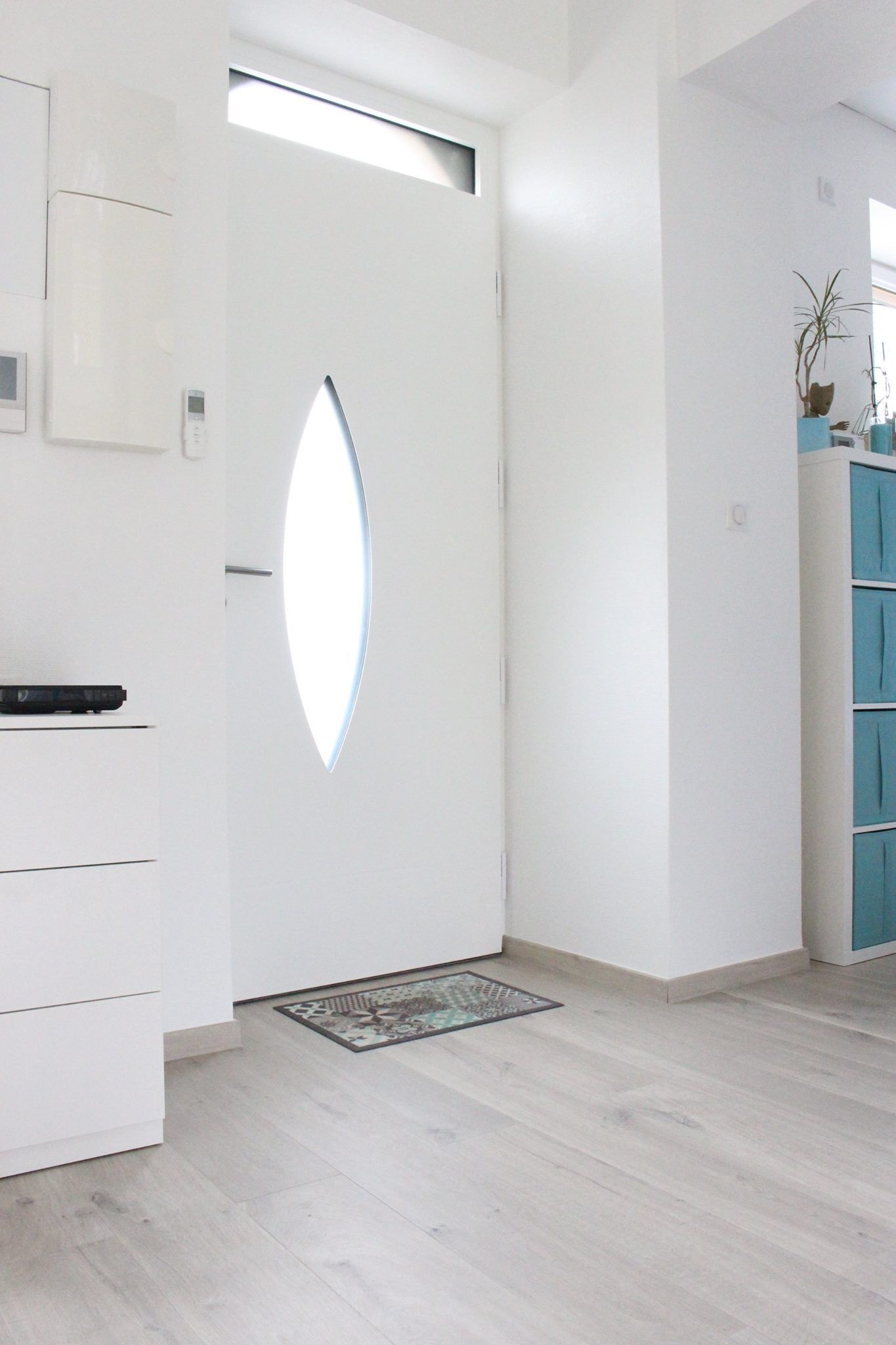 Extérieur d'une maison beige clair avec des boiseries et une porte d'entrée grises. Des palmiers en pot encadrent la porte et une chaise blanche se trouve à proximité.
