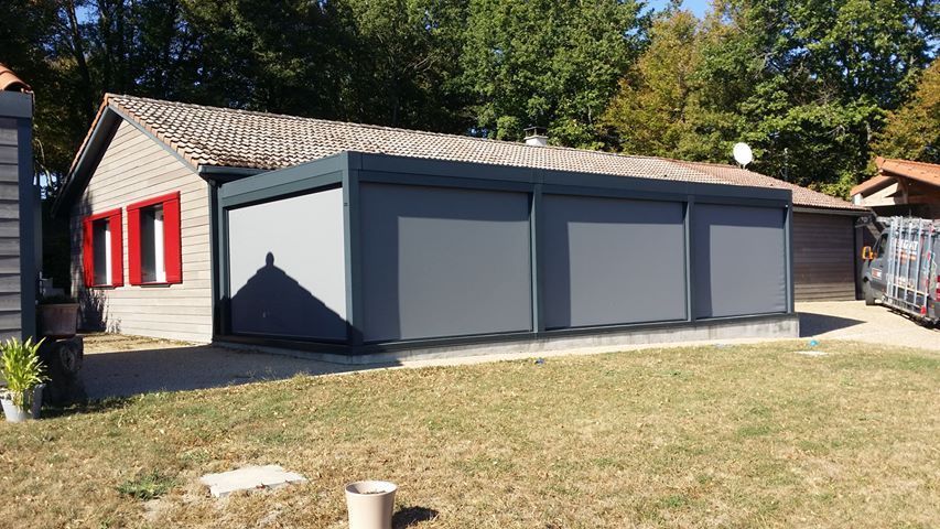 Une véranda aux murs gris, équipée de trois grands écrans rétractables, est attenante à une maison en briques aux volets rouges.