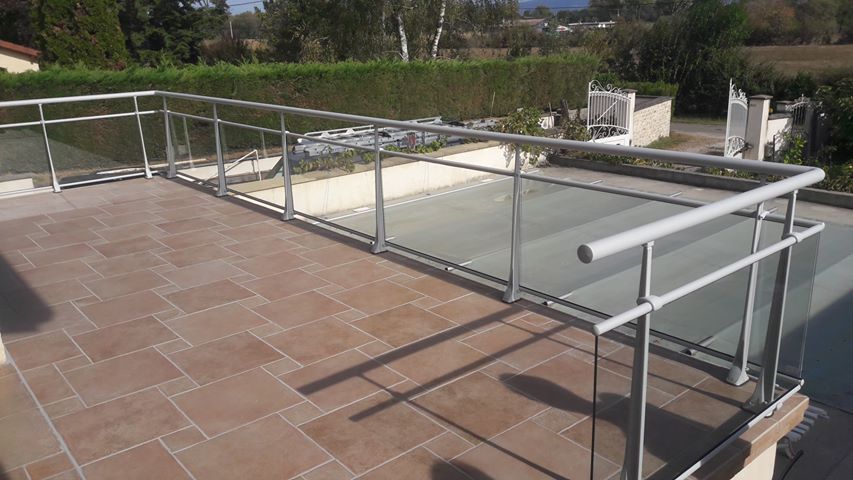 Balcon avec rambarde en verre et métal, donnant sur une végétation luxuriante et un ciel dégagé. Sol carrelé marron.