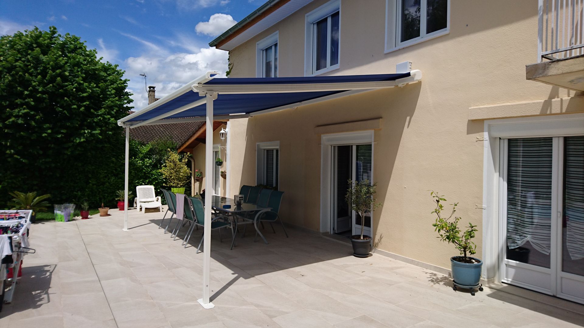Patio d'une maison avec un auvent, des sièges et des plantes en pot sous un ciel ensoleillé.