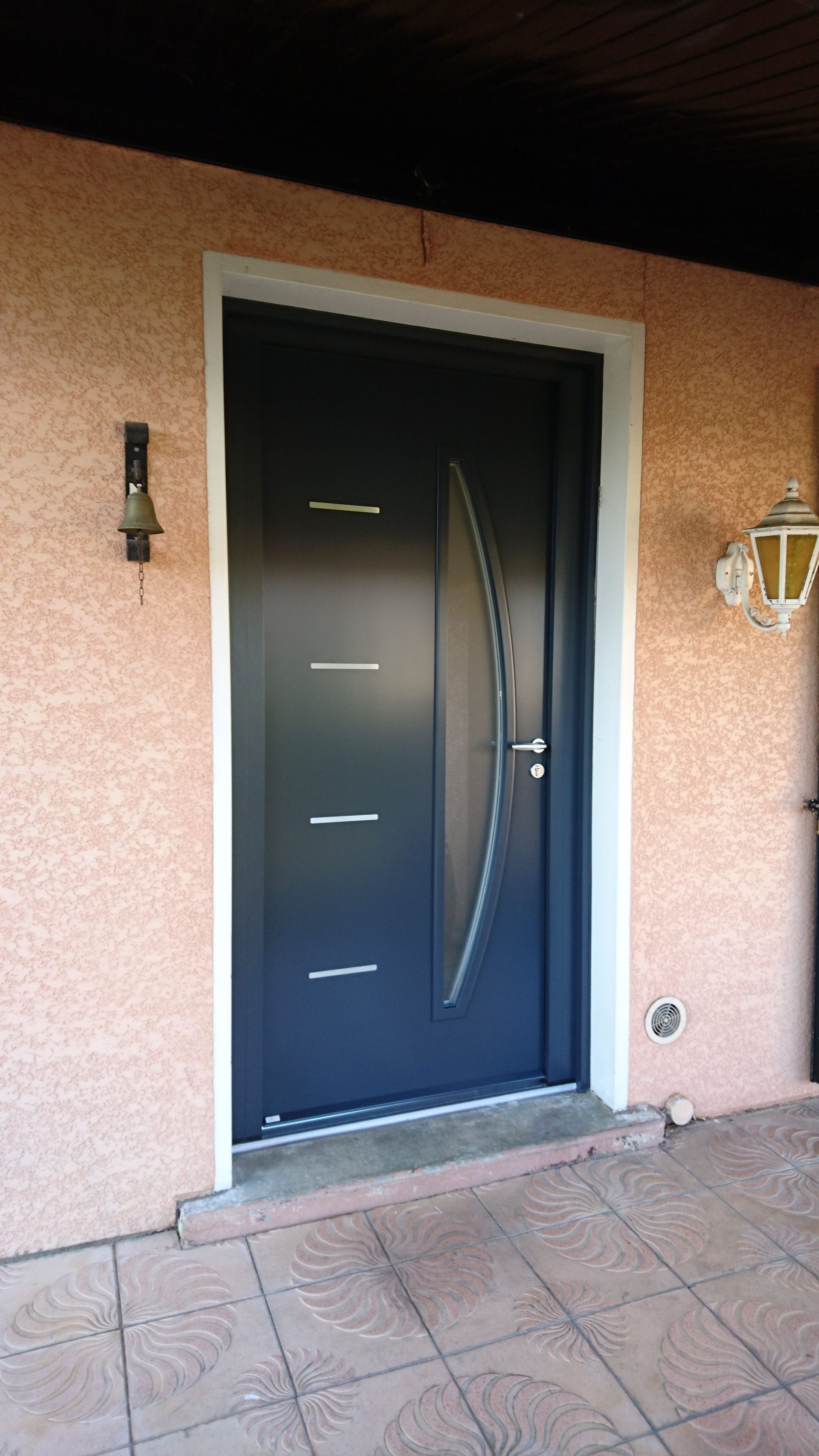 Porte d'entrée noire avec panneau de verre ovale et fentes horizontales, encadrement blanc sur un mur en stuc rose.