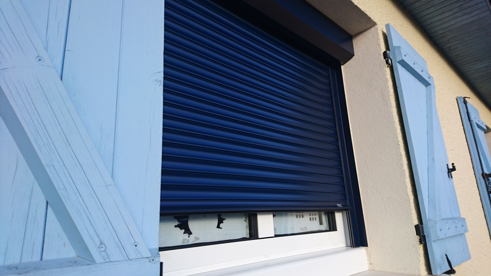 Volet bleu partiellement fermé sur une fenêtre à volets bleus.