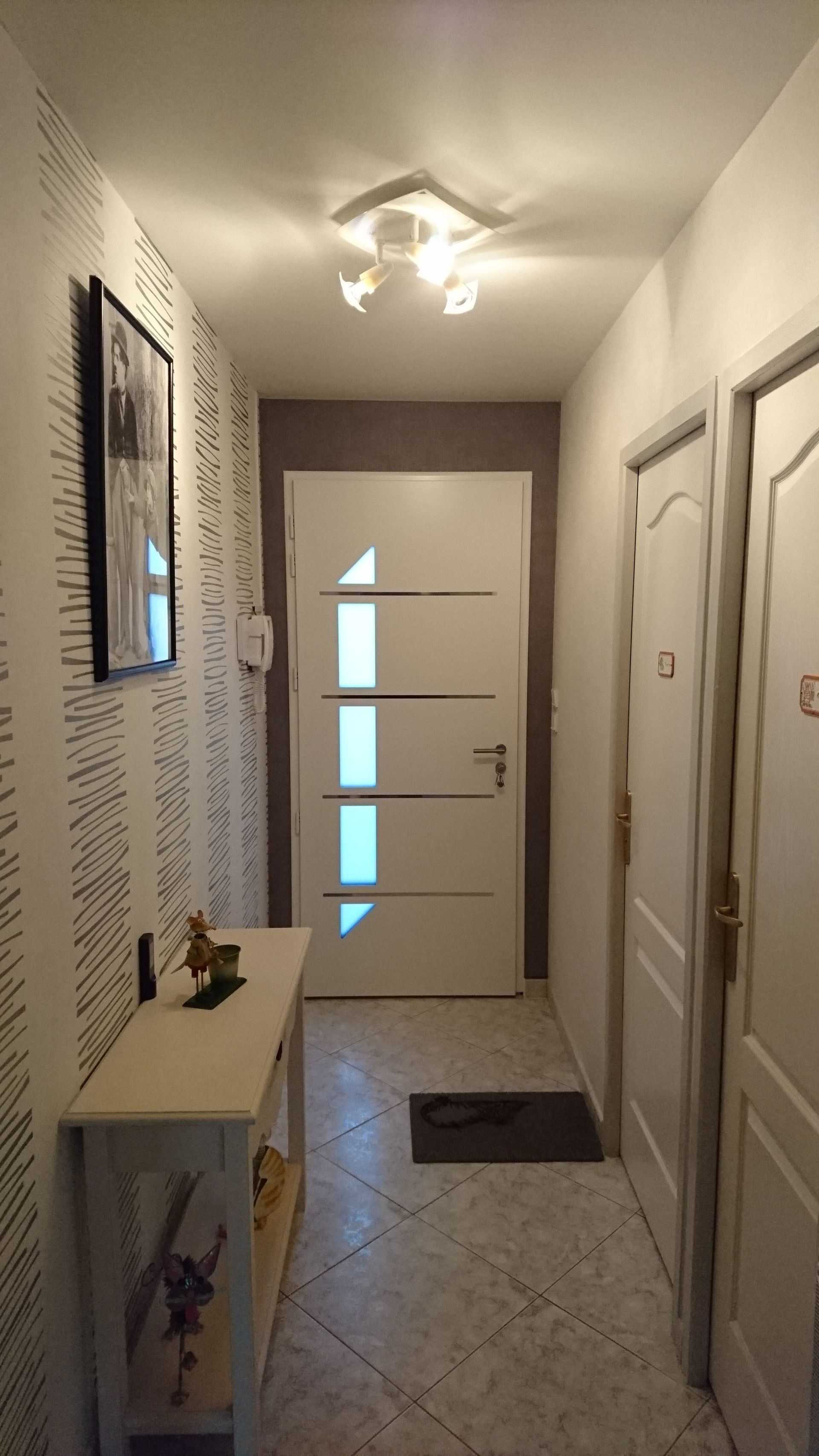 Couloir étroit avec portes blanches, console claire et porte d'entrée. Mur d'accent gris et papier peint à motifs.