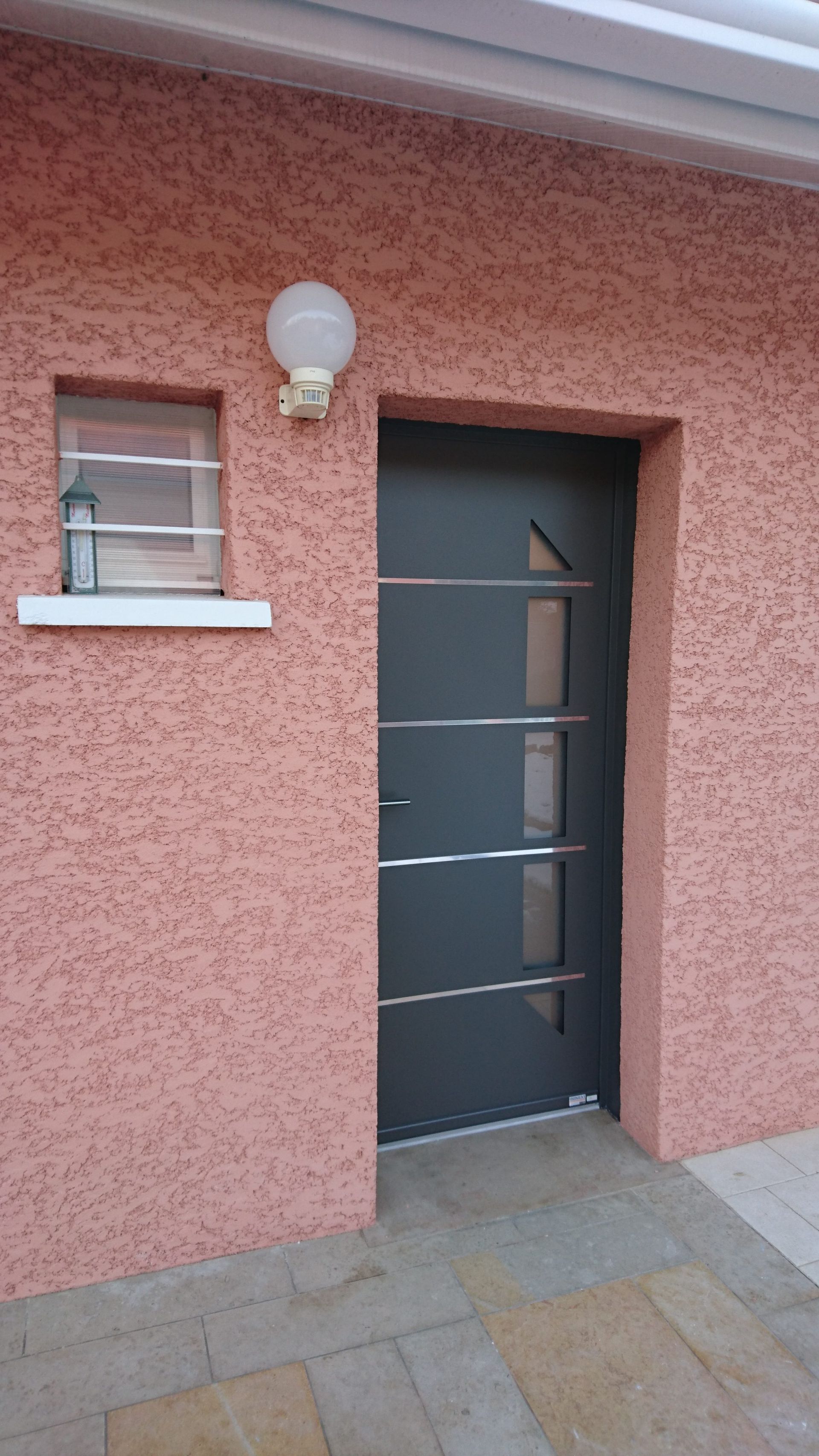 Porte grise sur un bâtiment couleur saumon, avec une petite fenêtre et un luminaire rond.