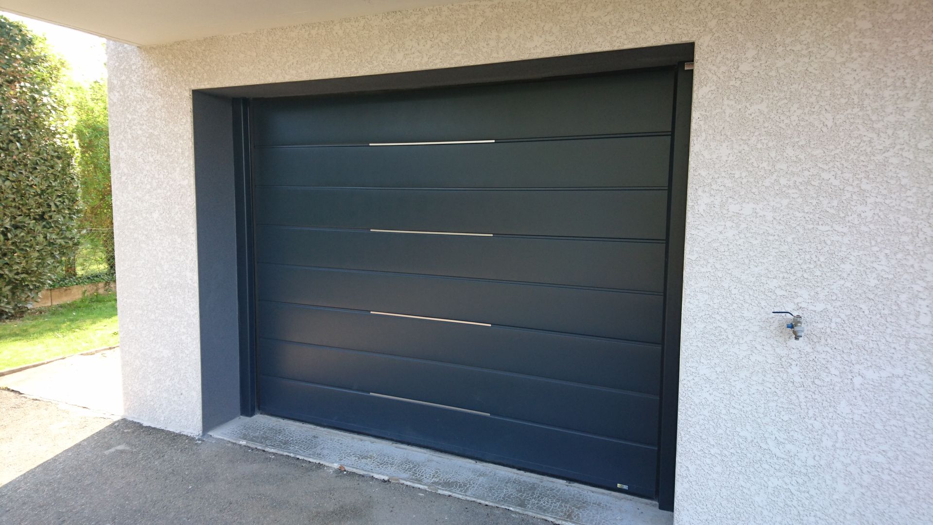 Porte de garage gris foncé à rayures horizontales, encastrée dans un mur en stuc gris clair.
