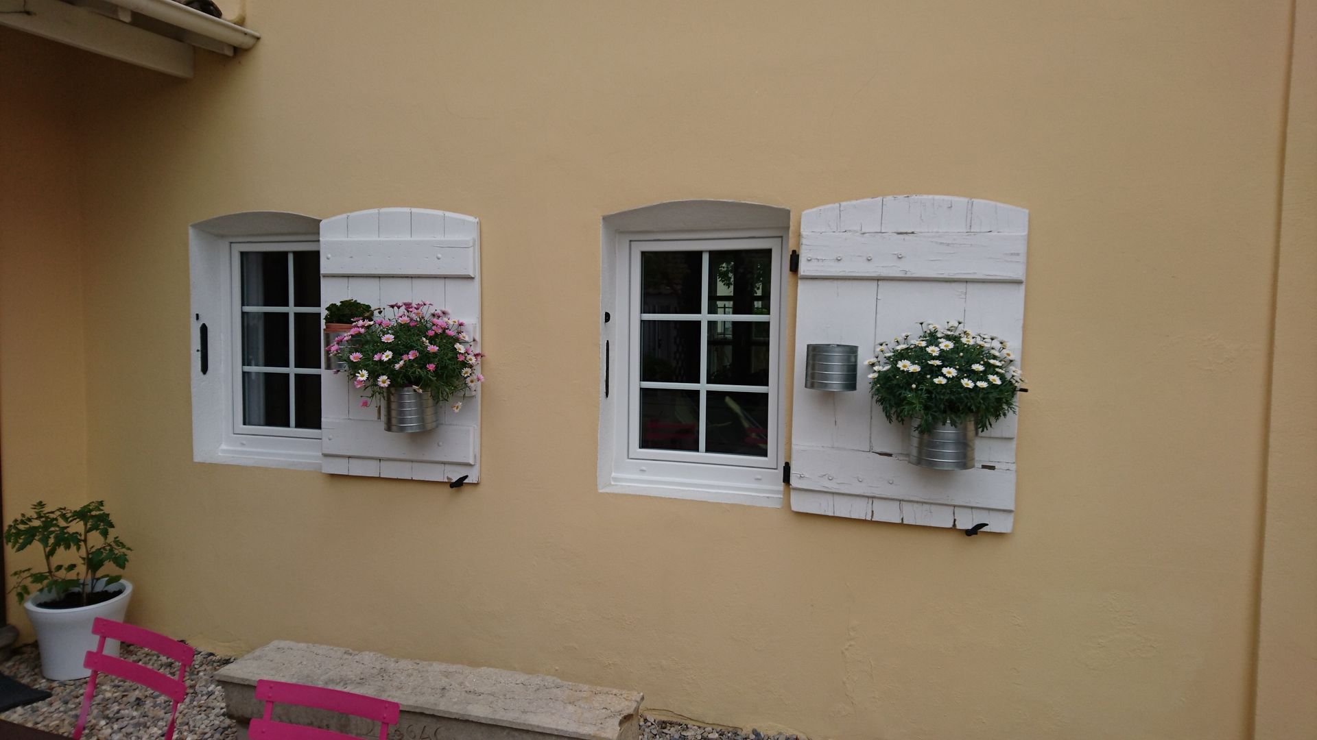 Deux fenêtres à cadre blanc avec volets blancs et jardinières sur un mur beige. Des chaises roses sont disposées devant.