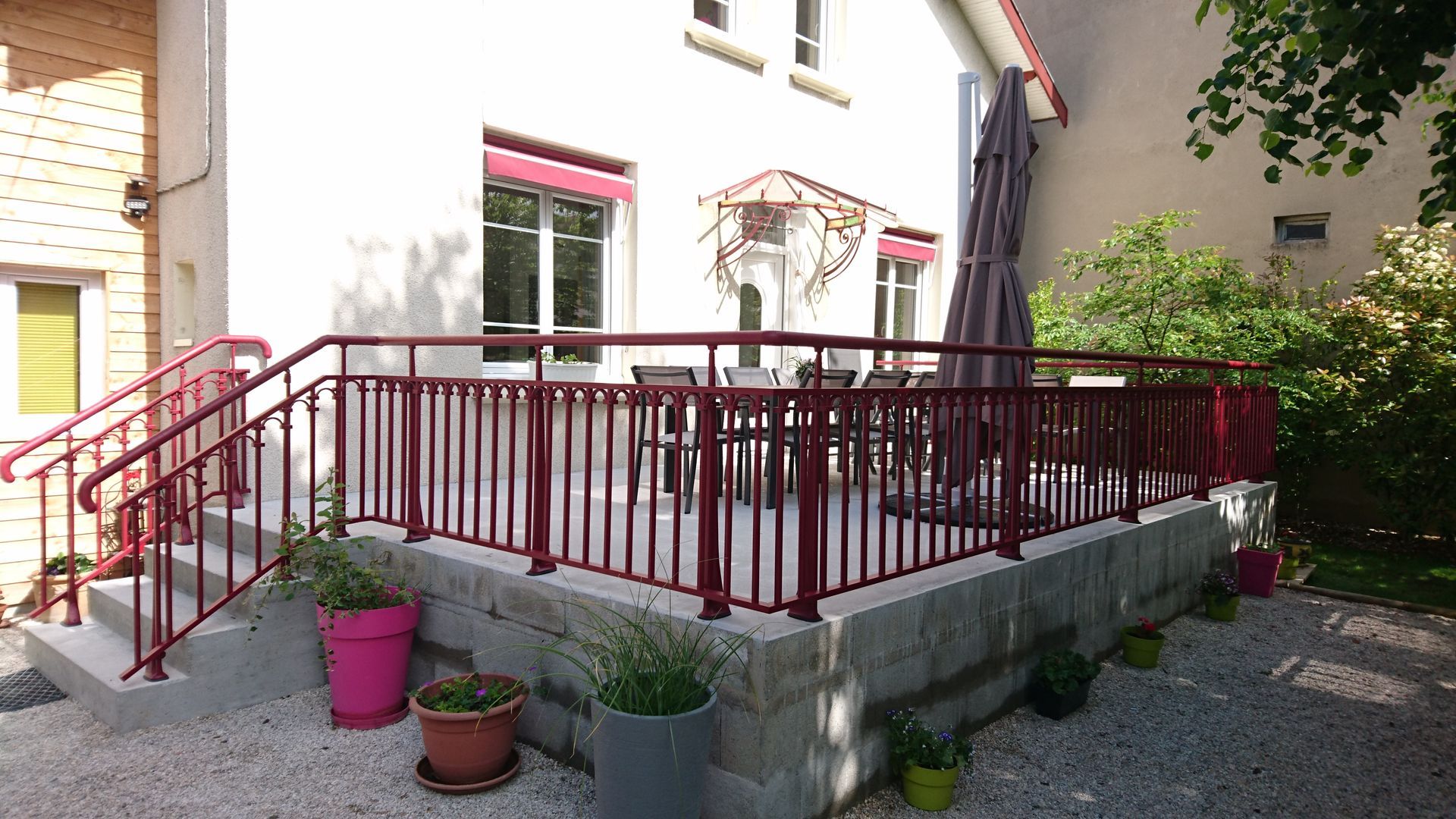 Une terrasse avec balustrade rouge et sièges extérieurs, attenante à un bâtiment de couleur claire, avec des plantes en pot devant.